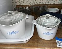 Two Corningware pots with glass lids, 2 quart and 1 quart, with blue cornflower design, placed on a white Corningware tray. Several glass bowls and ceramic ramekins visible in background.