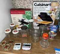Photo of kitchen items including Cuisinart waffle maker box, Pizzelle Baker cookie maker box, glass jars with floral lids, digital timers, jar lids, cups, vintage nut grinder, and sugar dispenser on kitchen counter.