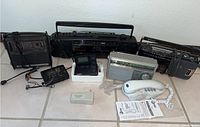 Lot of vintage electronics showing Emerson boombox, multiple radios including Panasonic and Sony, AT&T white phone, General Electric two-way radio, charger, and Sony Walkman on tile floor.