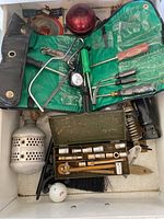 Top-down photo showing various small tools including a socket set in metal box, screwdrivers with green and transparent handles in roll-up pouch, a hacksaw frame, a golf ball, vintage metal parts, and a brush, packed loosely inside a drawer.