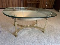 Oval glass table with clear glass top and brass base, shown standing on carpeted floor.