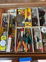 Wide view of two kitchen drawers containing assorted home repair tools and items such as pliers, screwdrivers, glues, tape measures, lightbulbs, keys, and padlocks, all neatly organized in compartments.