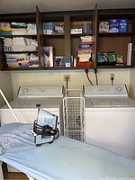 Photo of laundry room shelves with stacked towels and rags, Black & Decker iron on ironing board, heating pad, and washing machines in background.