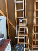 Full length view of ladders stacked in a garage corner including an aluminum extension ladder, 5-step wooden ladder with paint marks, and small folding stepstool in front.