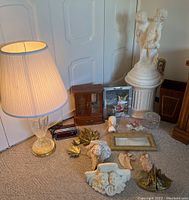 Glass table lamp with brass base and fabric shade, various angel statues, jewelry box, alarm clock, framed art, glass jar with lid, butterfly bookends, small decor items on carpeted floor.