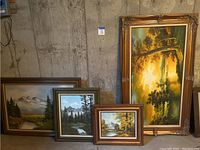 Four framed landscape paintings displayed against a concrete wall in a basement. Varied frame styles and sizes visible.