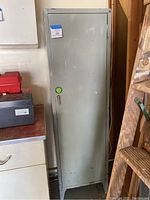 Front view of tall closed gray metal cabinet with auction tag labeled '88 metal cabinet'. Cabinet door is locked with a key in the lock.