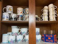 Cabinet shelf with assorted collectible mugs showing cityscape and animal designs and floral mugs stacked below them