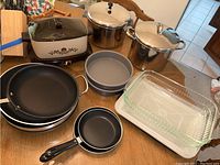 View of all kitchen cookware and appliances on table: slow cooker, frying pans, stockpots, pie dishes, casserole dishes.
