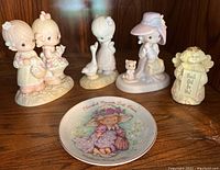 Lot setup with four Precious Moments figurines and one commemorative plate displayed on a wooden surface.