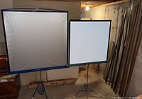 Two Da-Lite vintage projection screens on tripod stands and various wood trim pieces stored upright in the basement.