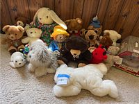 Group of various stuffed animals including Paddington Bear, Petster in box, and multiple bears, rabbit, dog, seal, and bug plushes displayed on carpeted floor