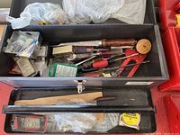Open large metal toolbox showing assorted hand tools, including hand saw, multiple screwdrivers with wood and plastic handles, measuring tape, and other small tools and hardware.