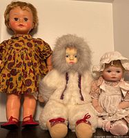 Group photo of the three dolls: vintage doll in floral dress, Eskimo doll in white furry outfit, and ceramic baby doll in pink and white dress.