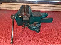 Metal surface mounted swivel vise placed on a red cloth showing the overall shape, swivel base, tightening handle, and jaw faces.