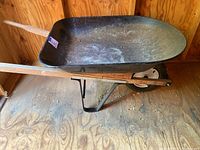 Side view of metal wheelbarrow showing black painted metal tray with rust and wear on inner surface, wooden handles, and single rubber tire.