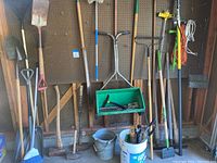 Long view of various shovels, rakes, and sticks against pegboard wall with buckets below