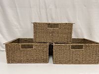 Three rectangular woven wicker storage bins displayed. Small placed on top of two larger bins side by side showing front view with built-in woven handles.