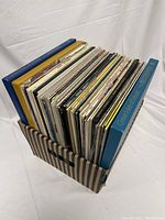Over 45 mixed vinyl records stored upright in a striped cardboard box, showing a variety of colorful record sleeves and boxed sets.