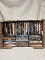 Top view of the open cardboard box filled with DVDs arranged vertically and stacked in two rows. Visible titles include 'Bugsy', 'The Switch', 'Pablo Escobar', 'American Beauty', 'G.I. Jane', among others.