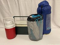 Photo showing all four items: red and white Coleman jug, green and white Coleman cooler, MEC Little Dipper child's sleeping bag in black and turquoise sack, and blue Bass Pro Shop youth sleeping bag in blue sack.