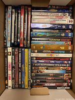 Overhead photo showing varied DVD and Blu-ray cases arranged in a box featuring movies and TV shows across genres including The Dark Knight, Whip It, The Fifth Element, Sherlock Holmes, and more.