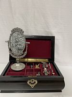 Black wooden jewelry box open showing red velvet interior containing rings and other jewelry with ornate oval mirror