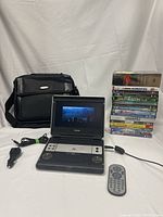 Overall lot showing Philips portable DVD player open with playing screen, black carrying case, remote control, car adapter, and stack of DVDs.