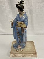 Front view of porcelain Japanese woman figurine in blue kimono holding tea set on maroon tray, standing on beige rectangular plate base.