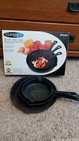 Set of three Heuck cast iron skillets displayed with original packaging box in the background.