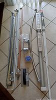 Five various curtain rods including one new in original plastic package, placed on tile floor alongside other construction items.