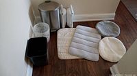 Full lot showing bathmats, toilet seat covers, toilet wands with holders, and three trashcans placed on the floor against a beige wall and hardwood floor.