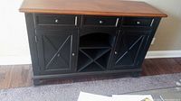 Front view of wood buffet table showing 3 upper drawers, 2 lower cabinets with X-braced door detailing, open central shelf with removable wine rack beneath, and brown top with black base.