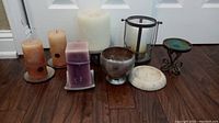 Photo showing multiple candles and holders arranged on floor including silver and ceramic holders, pillar candles, and several unique candle holders.