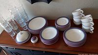 Photo of entire dishware and glassware set displayed on table, showing plates, bowls, cups, saucers, butter dish, salt shaker, and clear glass tumblers in two sizes