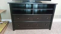 Front view of dark brown/black wooden entertainment console with four drawers below and four open shelves above, standing on carpeted floor.