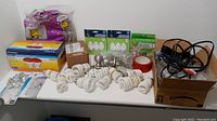 Various bulbs (infrared, halogen MR16 round, halogen T2.5), numerous compact fluorescent bulbs, adhesive hooks, red tape roll, socket sealers, and box of assorted cords arranged on a white table.