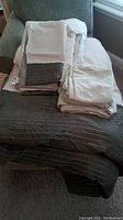 All bedding items stacked on chair showing variety in colors and textures, including beige and dark sage microfiber pieces and beige sheets.