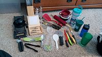 Overview of many kitchen utensils and beverage containers on beige carpet, including knives, grinders, mini spice jar, spatula, green cup, and plastic tumblers.