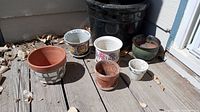 Seven assorted plant pots arranged on a deck, including terracotta, ceramic, and plastic pots