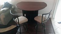 Round counter height table with fold-down sides and storage below, dark wood finish, and two beige cushioned counter height stools.