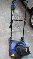 Front view of Snowjoe snow thrower showing blue plastic motor housing, black chute, metal handle with blue grip and control cables.