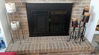 Overall view showing silver plant stand holding two large used cream/white candles on left and three black wrought metal floor candlesticks with tri-color used candles on the right, placed on a brick hearth in front of a closed fireplace.