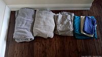 Full lot of folded bedding items arranged on a wood floor, including grey blankets, sheets in grey and teal colors, and multiple pillowcases in various colors