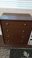 Brown pressed board chest with four drawers, round metal knobs, wood grain finish, minor damage not visible, placed indoors on carpet.