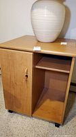 Front and top view of light wood finish locking storage cabinet with a key in the door lock, placed on carpeted floor with a vase on top.