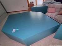 Photo showing the teal polygonal coffee table with smooth finish and solid structure in a living room setting next to a sofa.