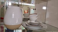 Photo showing a large pottery pitcher with a white glazed upper part and unglazed brown bottom next to stacked Rosenthal Kontrollstelle china plates and bowls with brown rim accents.