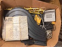 Contents of cardboard box showing black plastic curved track sections, yellow guardrails, yellow and black controllers, metal power supply unit and instruction sheet labeled Aurora AFX Model Motoring.