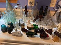 Overview of various clear and colored glass bottles, Avon collectible decanters shaped as vehicles and barrels, green glass insulators, and some ceramic busts on a kitchen counter.
